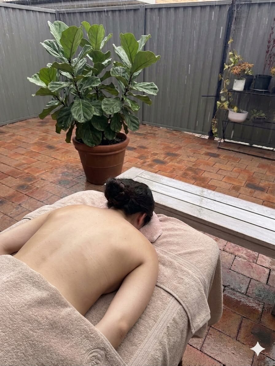 Kerry giving a remedial back massage on a padded table in a leafy courtyard, beside a large fiddle-leaf fig